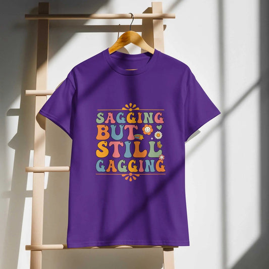 Purple Saggin Still Gaggin DryBlend® T-Shirt hanging on wooden rack with colorful text design.