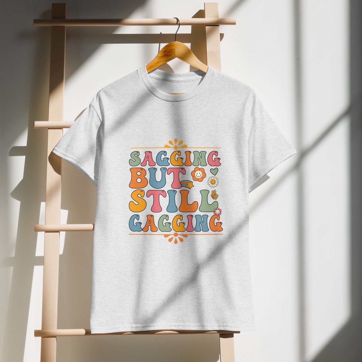 Saggin Still Gaggin DryBlend® T-Shirt with colorful text design hanging on wooden rack.