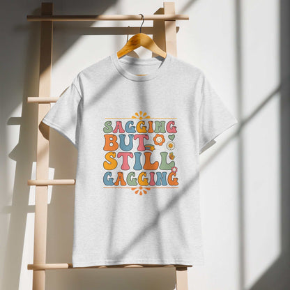 Saggin Still Gaggin DryBlend® T-Shirt with colorful text design hanging on wooden rack.