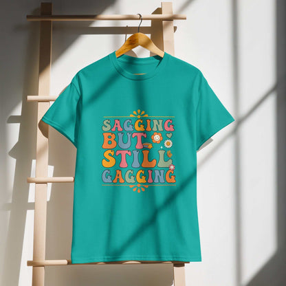 Teal Saggin Still Gaggin DryBlend® T-shirt with colorful text, hanging on a wooden stand in sunlight.
