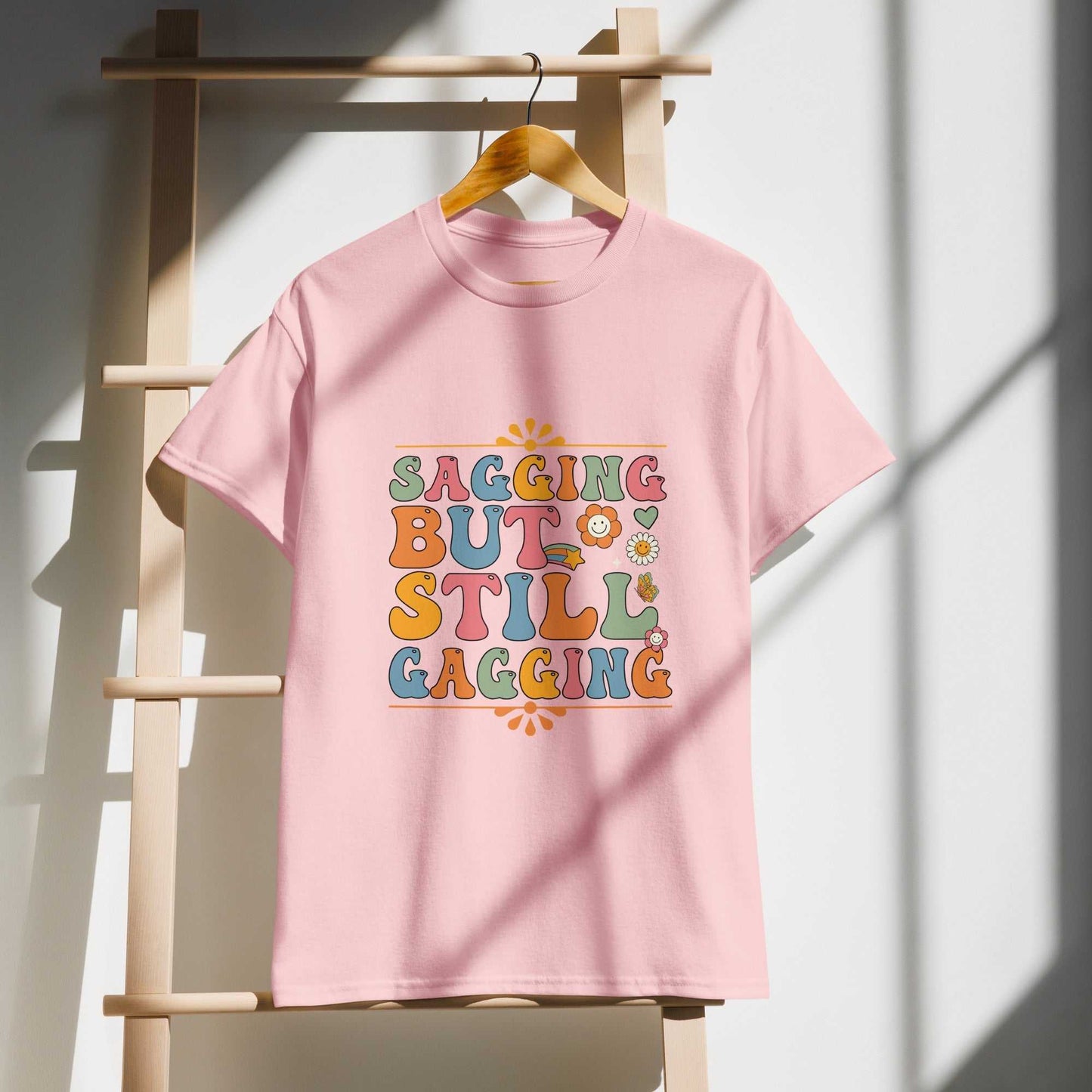 Saggin Still Gaggin DryBlend T-Shirt in pink hanging on ladder, featuring colorful text design.