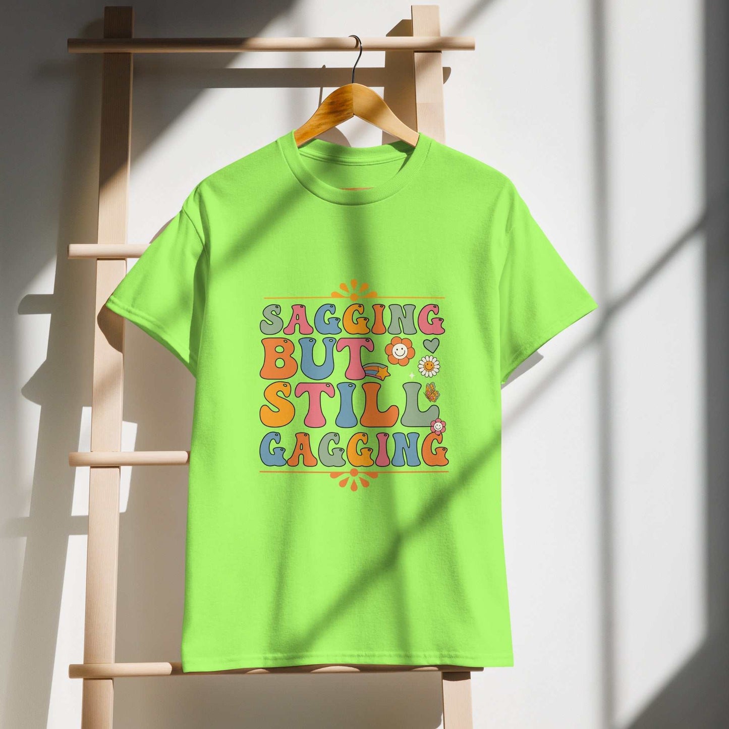 Saggin Still Gaggin DryBlend T-Shirt hanging on a wooden rack, featuring colorful text design on a green fabric with sunlight casting shadows.